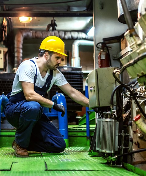 Diesel Mechanic in Overalls and Helmet Kneeling Inside Ship and Repairing Engine